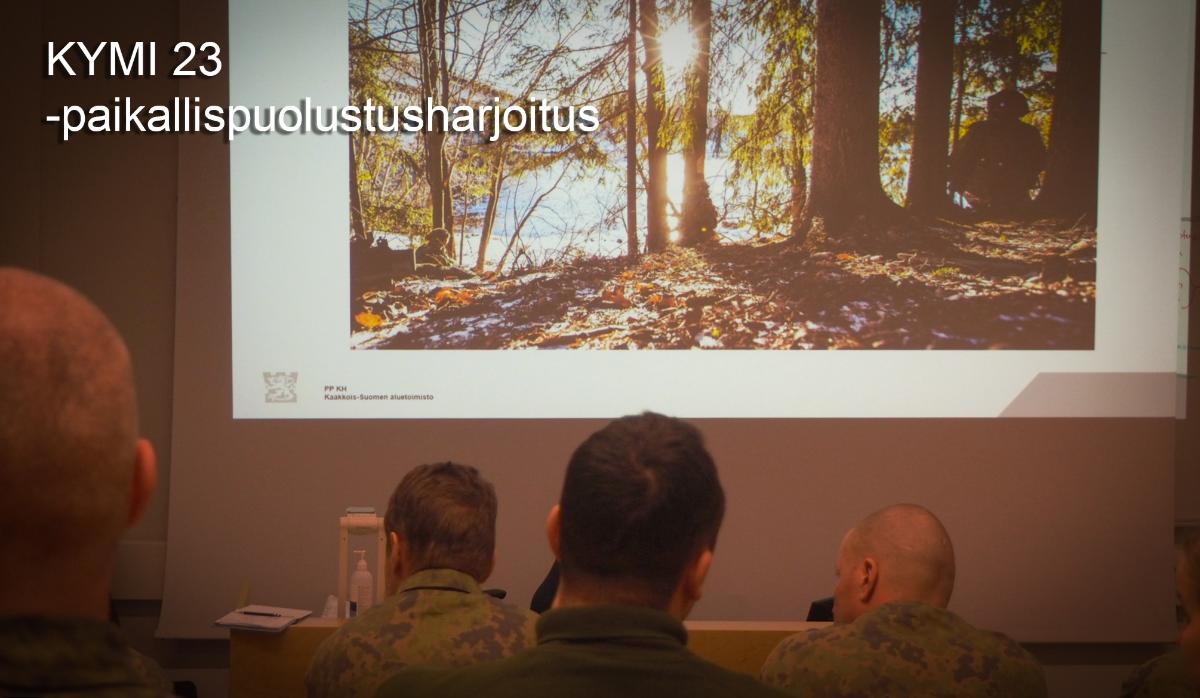 Maastopukuisia henkilöitä sisätiloissa katsomassa valkokankaalla olevaa kuvaa.